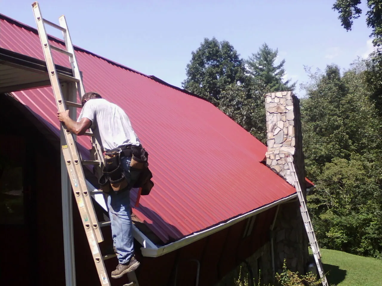 Roofer on a ladder beside a red metal roof and stone chimney — Soffit & Fascia Repair by Roofing Brothers in North Waterboro
